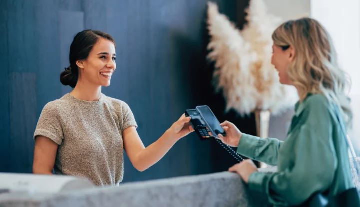 Maximize hotel revenue with e-Commerce solutions: Boost booking spend - Woman taps phone to pay at hotel front desk - Sabre Hospitality