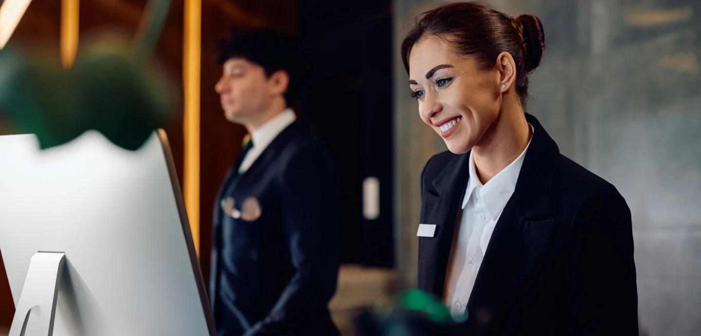 A woman smiles at a front desk at a hotel, looking at her computer. Top 5 things to look for in hotel management software - Sabre Hospitality